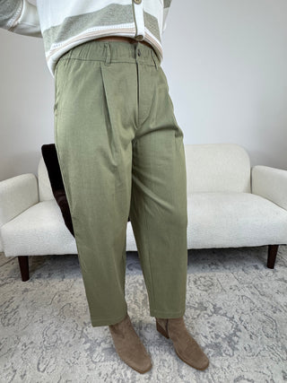A person wearing olive green pleated trousers, styled with a striped cardigan and brown suede boots, standing in a living room setting with a white textured sofa and patterned rug.