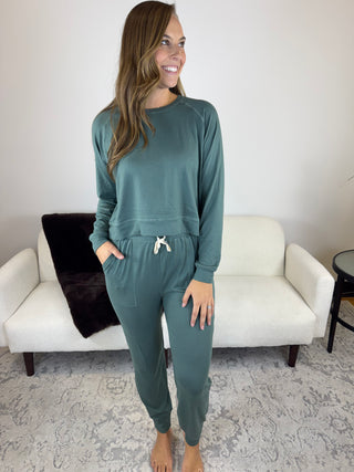 A woman modeling a matching two-piece lounge set in a sage green color, consisting of a long-sleeve sweatshirt and jogger pants with a white drawstring, standing in a living room setting.