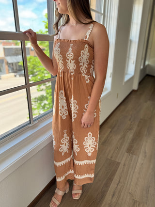 Woman wearing a brown dress with white lace patterns standing by a window.