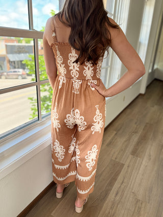 Person wearing a brown jumpsuit with white lace patterns standing by a window.