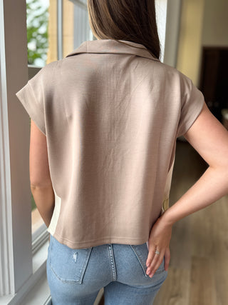 Person wearing a beige sleeveless top and blue jeans standing by a window.