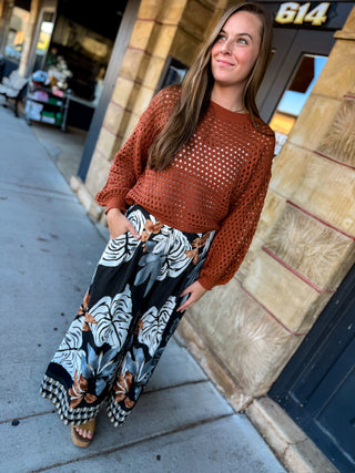 A woman wearing a rust-orange long-sleeve crochet knit top and black wide-leg pants with a white, gray, and brown tropical floral print, standing on a city sidewalk.