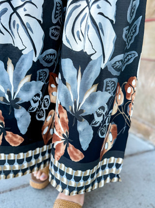Close-up of wide-leg pants with a bold botanical print in navy, light gray, white, and terracotta, worn with woven sandals on a sidewalk.