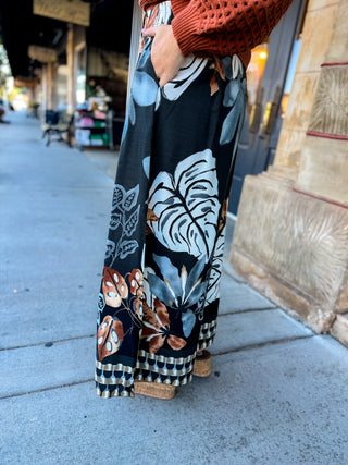 A person standing on a city sidewalk wearing wide-leg maxi pants with a tropical leaf and floral print in black, beige, brown, and blue-gray, paired with a rust-orange knit top.