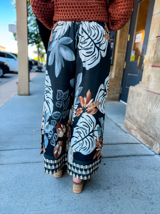A person standing outdoors on a sidewalk wearing wide-leg cropped pants with a tropical leaf print in navy, white, and rust tones, paired with a rust-colored knit top and cork wedge sandals.