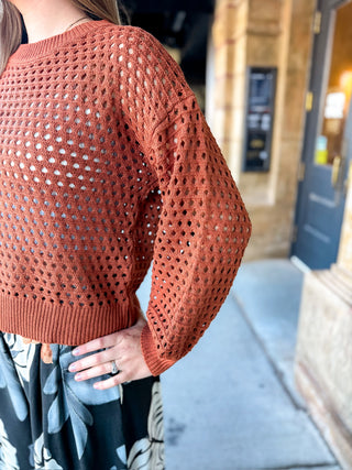 A person wearing a rust-orange open-knit long-sleeve sweater, paired with a black and white floral skirt, standing outdoors on a sidewalk.