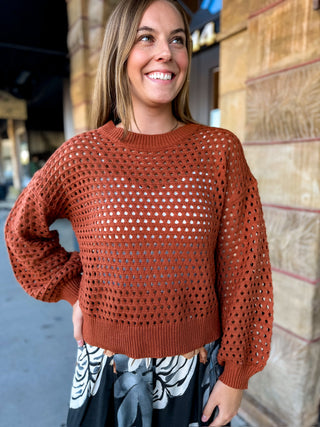 A woman wearing a rust-orange open-knit crewneck sweater with balloon sleeves, standing outdoors against a stone wall.