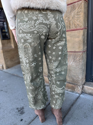A person shown from the back wearing olive green wide-leg pants with a white paisley and floral pattern, standing on a sidewalk next to a stone building.