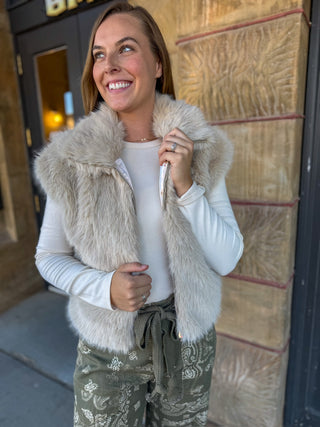 A woman smiling and looking upward while wearing a light beige faux fur vest over a white long-sleeve top and olive green patterned pants, standing outdoors in front of a stone wall.
