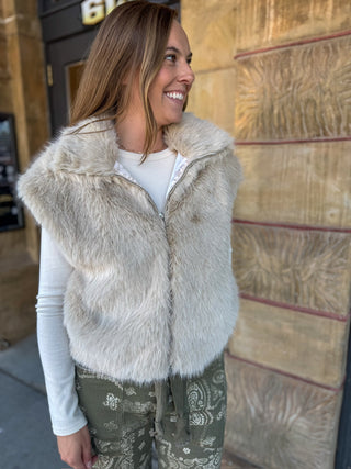 A woman wearing a light beige faux fur zip-up vest over a white long-sleeve shirt and olive green paisley pants, standing outdoors against a stone wall.