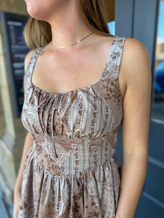 A woman wearing a sleeveless midi dress with a brown and beige paisley pattern, featuring a gathered bodice and thin straps.
