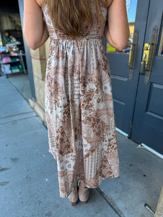A woman viewed from behind wearing a sleeveless midi dress with a brown and beige paisley pattern, standing on a sidewalk in front of blue-gray doors.