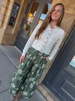 A woman wearing a white textured button-up cardigan and a green wide-leg skirt with a white floral vine pattern, standing on a sidewalk in front of a building with dark doors.
