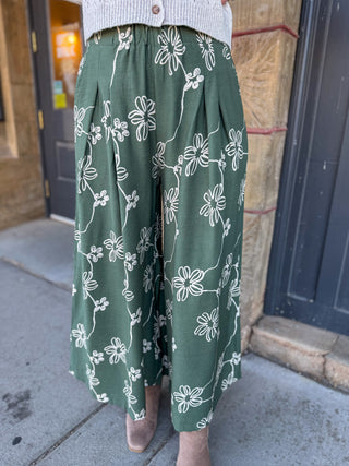 A person wearing wide-leg cropped pants in sage green with a white floral print, paired with a light gray knit top and beige shoes, standing on a sidewalk in front of a building.