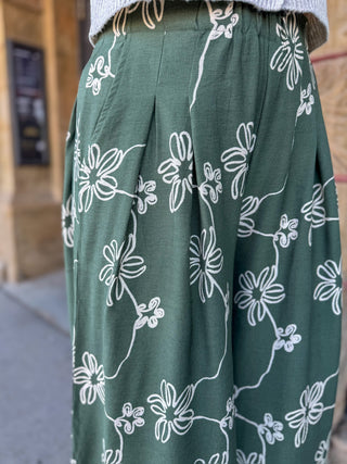 A close-up of a person wearing wide-leg pleated green trousers with a white floral line-art pattern, paired with a light gray ribbed top. The background is a softly blurred outdoor street scene.