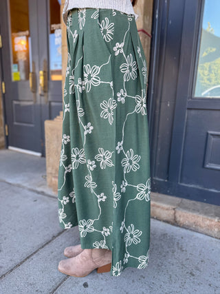A person wearing a long green maxi skirt with a white floral line-art pattern, paired with beige ankle boots, standing on a sidewalk in front of dark blue doors.