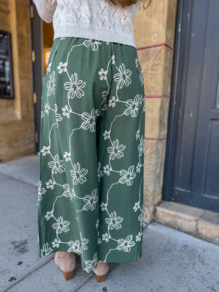 A person wearing wide-leg green pants with a white floral vine pattern, paired with a white knit sweater and beige heels, standing on a sidewalk in front of a building.