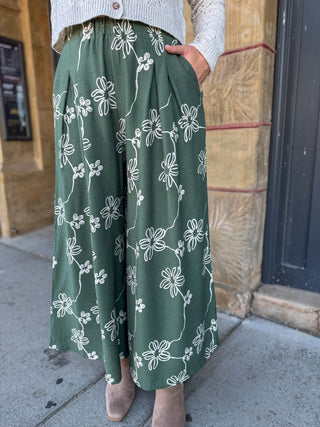 A person wearing a pair of wide-leg green pants with a white floral line-art print, paired with a light gray knit top and beige ankle boots, standing outdoors on a sidewalk.
