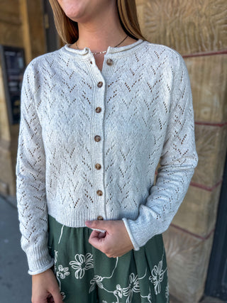A woman wearing a light cream open-knit button-front cardigan with brown buttons, paired with a green floral skirt, standing outdoors against a stone wall.