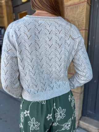A person shown from the back wearing a light gray open-knit long-sleeve sweater and green floral pants, standing outdoors against a stone wall and dark door.