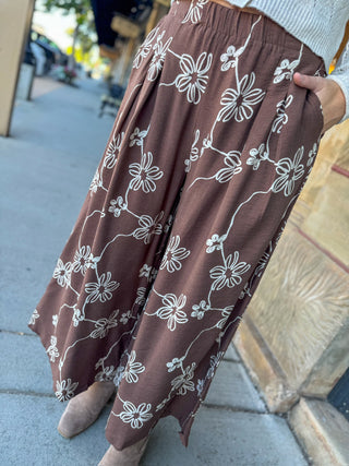 A person wearing a brown wide-leg skirt with a white floral line pattern, standing outdoors on a sidewalk next to a stone wall.