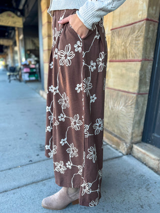 A person wearing brown wide-leg pants with a white floral print, standing on a city sidewalk next to a stone building.