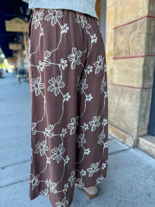 A person wearing wide-leg brown pants with a white floral vine pattern, standing outdoors on a sidewalk next to a stone building.