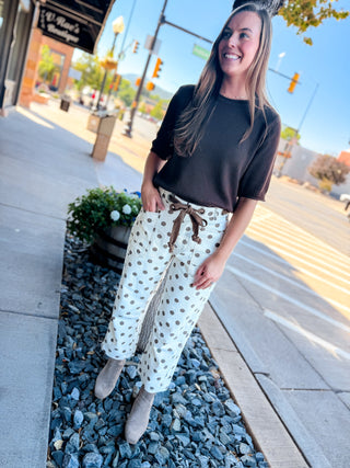 A woman stands on a city sidewalk wearing a dark brown short-sleeve top and white drawstring pants with brown polka dots, paired with taupe ankle boots.
