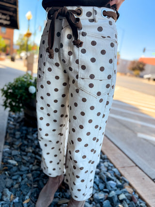 A person wearing cream-colored cropped pants with a brown polka dot pattern, a brown drawstring waist, and a matching brown belt, standing outdoors on a sidewalk.