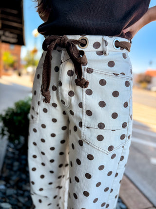 A close-up of a person wearing cream-colored pants with dark brown polka dots and a matching brown drawstring belt, standing outdoors on a sunny day.