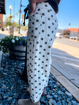 White denim pants with a dark brown polka dot pattern, worn by a person standing outdoors on a gravel path next to a street.