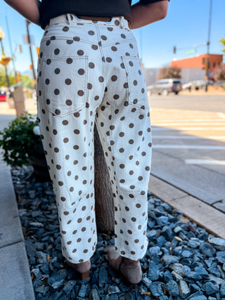 White denim pants with a brown polka dot pattern, shown from the back on a person standing outdoors on a gravel path near a street.