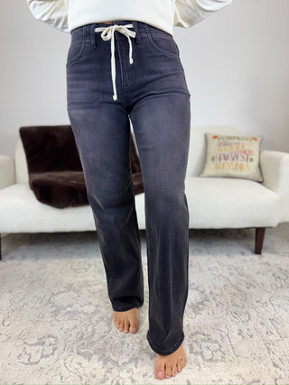 A person wearing dark charcoal gray drawstring jeans, standing barefoot on a patterned rug in a living room setting.