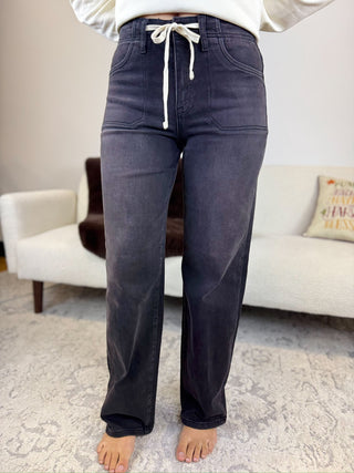 A person wearing dark charcoal gray drawstring jeans, standing barefoot on a patterned rug in a living room setting.