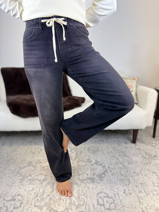 A person wearing dark navy blue wide-leg jeans with a white drawstring waist, standing barefoot on a patterned rug in a living room setting.