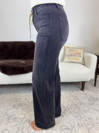 A person shown from the waist down wearing dark charcoal gray wide-leg jeans in a living room setting with a white armchair and patterned rug.