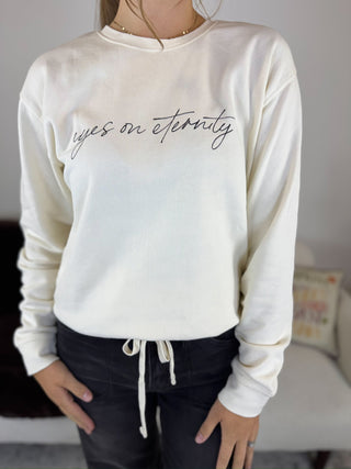 A person wearing a cream-colored long-sleeve sweatshirt with 'eyes on eternity' written across the chest in black script, paired with dark drawstring pants.