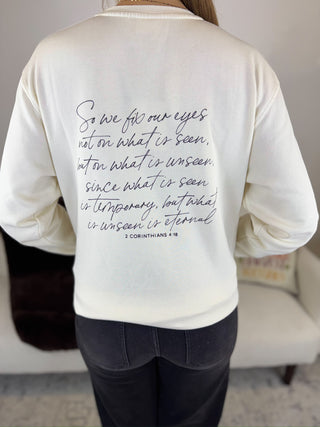 A person shown from the back wearing a cream-colored long-sleeve shirt with a black cursive Bible verse printed on the back, paired with dark gray jeans in an indoor setting.