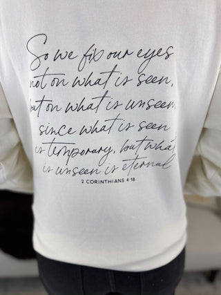 A white long-sleeve t-shirt featuring a black cursive hand-written style Bible verse from 2 Corinthians 4:18 printed on the back. The shirt is shown on a person wearing black pants.