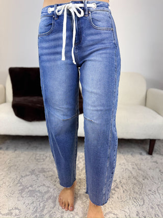 A person wearing blue denim jeans with a white drawstring waist, standing barefoot on a patterned rug in front of a light-colored sofa.