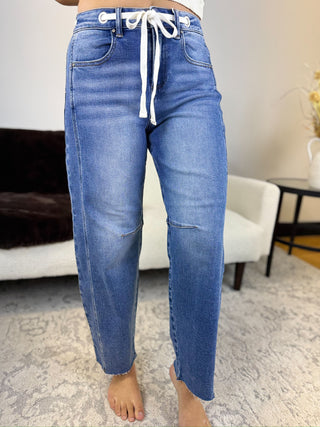 A person wearing blue denim jeans with a white drawstring waist, standing barefoot in a living room setting.