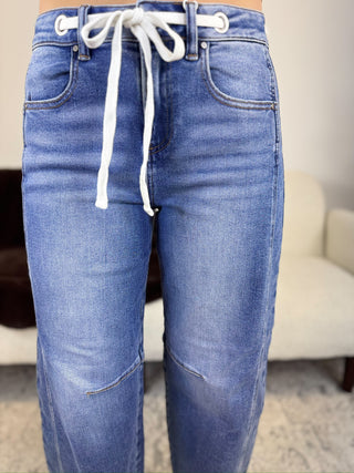 Close-up of a person wearing blue denim jeans with a white drawstring waist and white metal eyelets, standing indoors with a blurred chair and carpet in the background.