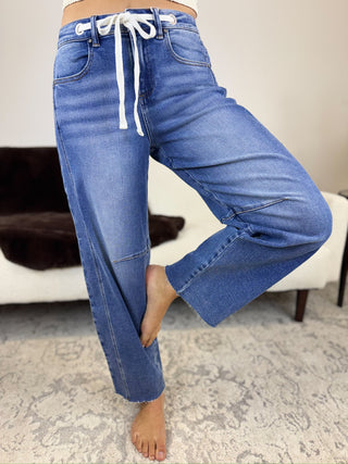 A person wearing blue wide-leg jeans with a white drawstring waist, standing barefoot on a patterned rug in an indoor room.
