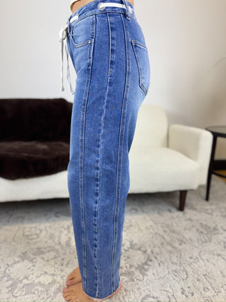 A person wearing blue denim jeans shown from the waist down in a side profile, standing barefoot in a living room with a white armchair and patterned rug in the background.