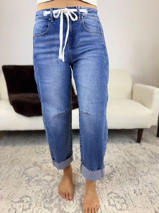 A person wearing blue denim jeans with a white drawstring waist and rolled cuffs, standing barefoot on a patterned rug in a living room setting.