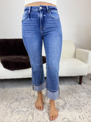 A person wearing medium-wash blue cropped jeans with rolled cuffs, standing barefoot on a patterned rug in front of a white sofa.