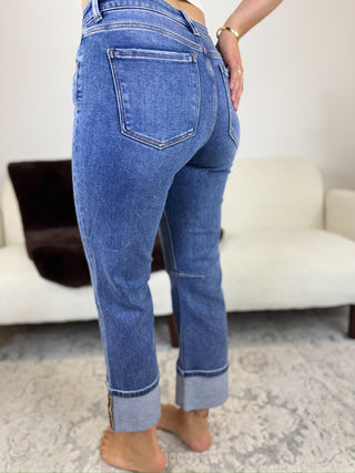 A person shown from the back wearing medium-wash blue cuffed jeans in a living room setting with a white sofa and patterned rug.