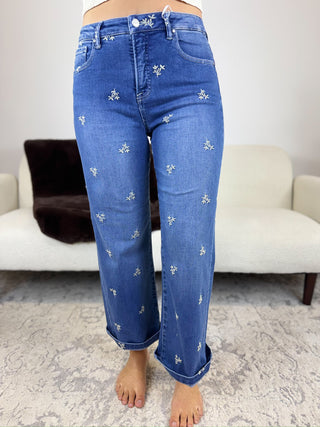 A person wearing blue denim jeans with small white embroidered floral motifs, shown from the waist down in a living room setting with a sofa and rug in the background.