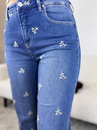 Close-up of a person wearing blue denim jeans with small white embroidered floral motifs scattered across the fabric, shown in a softly lit indoor setting.