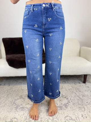 A person wearing blue denim jeans with small white embroidered floral details, shown from the waist down in a living room setting.